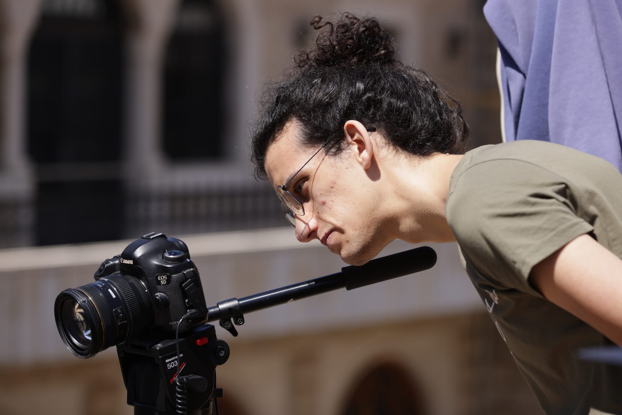 A videographer leans in to check the frame on a camera fitted with a shotgun microphone