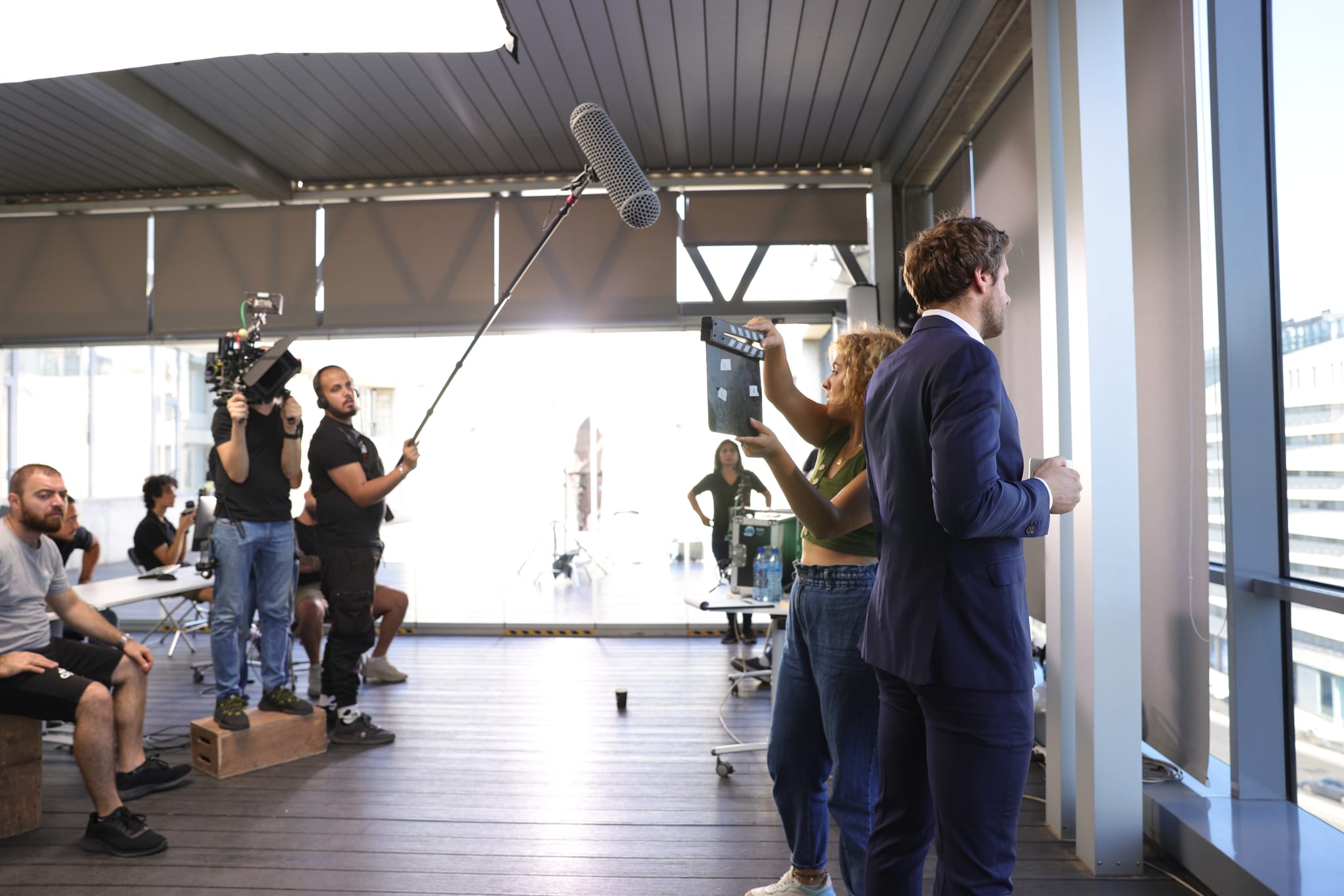 A boom operator and camera crew film a presenter in a suit inside a glass-walled office